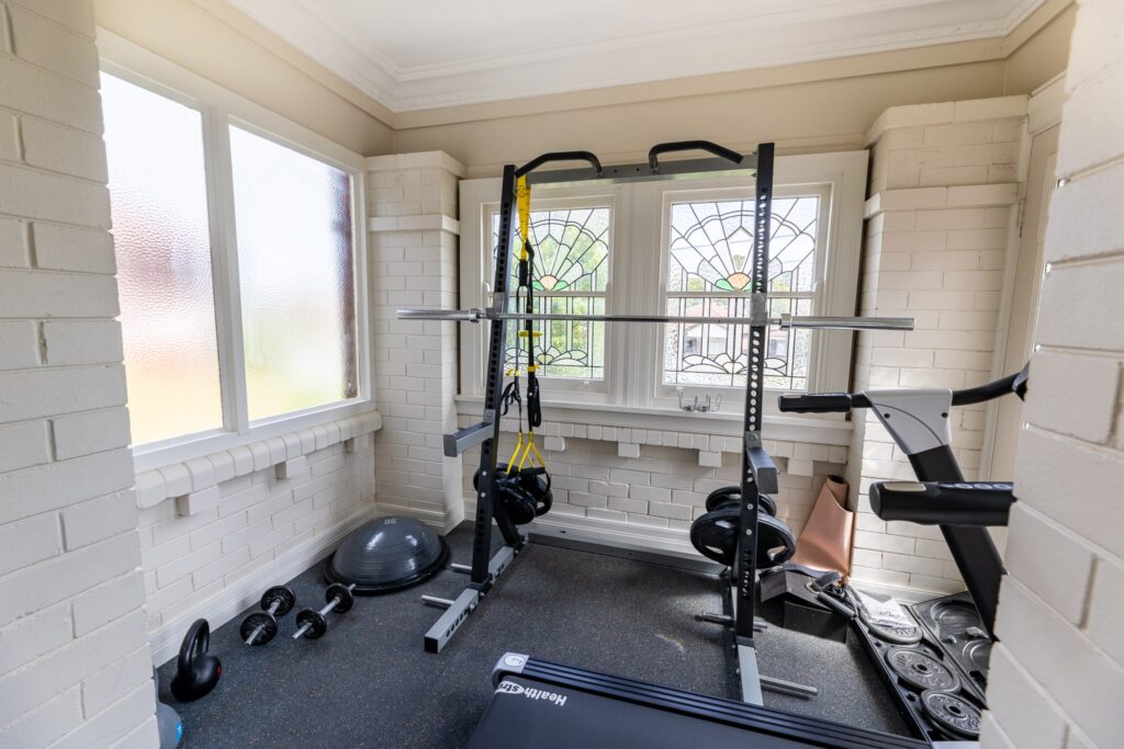 About us. Picture of functional movement gym area with equipment. A treadmill, free weights and a half rack. The room is cream in colour with beautiful lead light windows