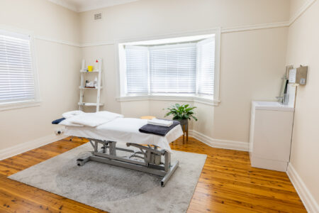 Picture of chiropractic treatment room. A chiropractic table is in the middle of the room on a blue rug. There is a desk and office chair in one corner and a chair for patients to sit on located in another. There is a bay window and the wall colour is cream. A white sink is located in a corner and a plant is located under the window. Remedial massage and pain therapy