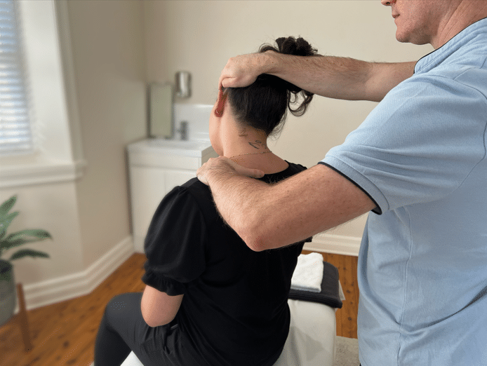 Five Dock Osteopathic and Chiropractic male chiropractor wearing a light blue shirt performing a stretch on the neck of a young female patient. The doctors right hand is placed on the head of the patient and the left hand on the shoulder of the female patient