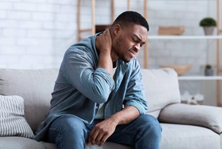 Middle aged African American man sitting on a grey lounge holding the back of his neck. He is wearing blue jeans and a blue and white checked shirt. He has a pained looked on his face. Auto related accidents can cause disc pain like whiplash.