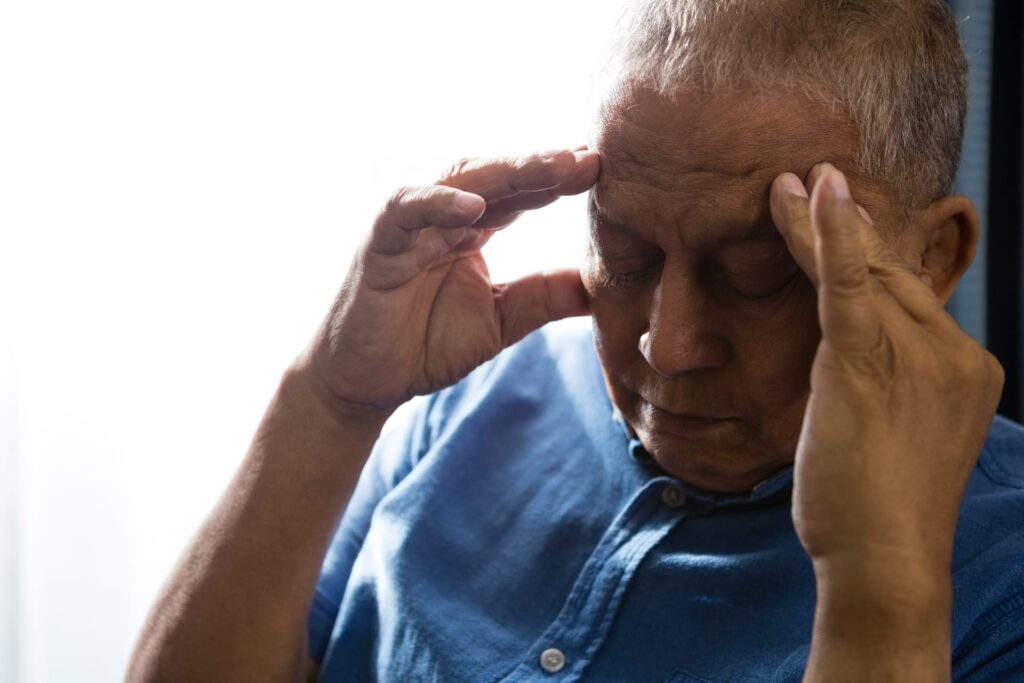 Elderly gentlemen holding both side of his head with his hands. He has a look of pain on his face. He is wearing blue Demin shirt.