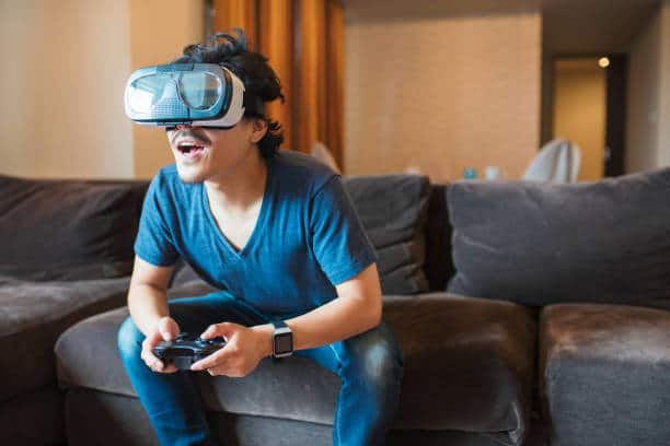 Young adult male using a virtual reality headset. Sitting on brown suede lounge. Wearing blue v neck T shirt and blue jeans. Sit leaning forward onto shoulders can damage rotator cuff.