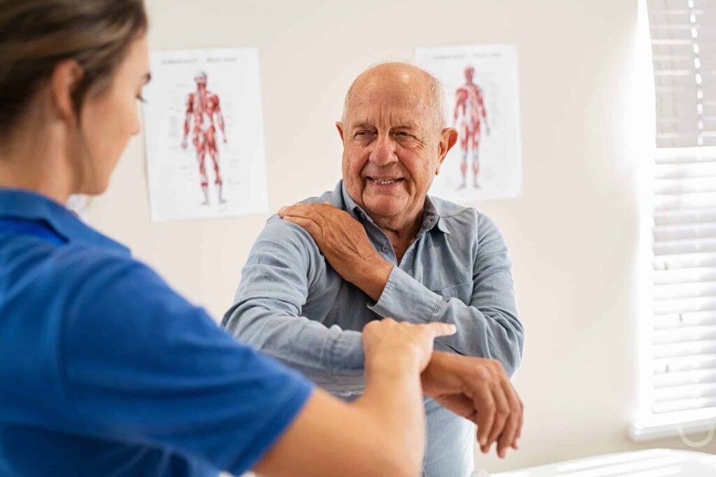 An elderly gentlemen is smiling as he shows a young female doctor where his right shoulder pain is coming from. His arm is at ninety degrees to his side. He is wearing a button up blue shirt. Anatomy poster are in the background on a cream coloured wall. Chiro Shoulder pain