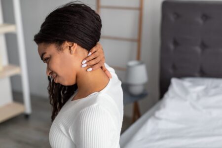 A young lady is sitting on the side of her bed wearing a white long sleeve top. She is reaching around behind her neck with her right hand as she lets her neck hang forward at 30 degrees. She has a look of discomfort on her face.