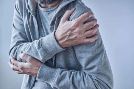A middle aged man with a grey beard is clutching his left shoulder in pain. He is wearing a grey lip up jumper and a grey shirt. He has a posture of bending forward at 15 degrees in shoulder pain. rotator cuff injury.