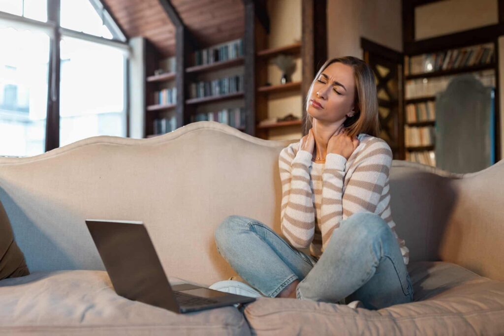 A young female is sitting on her lounge crossed legged facing the side arm rest. She is wearing blue jeans and a stripe brown and cream jumper. She has bookshelves behind her and a large window. She has a laptop computer in front of her. She is sitting holding her neck with a look of discomfort. She is bending her neck to the left as she sits.