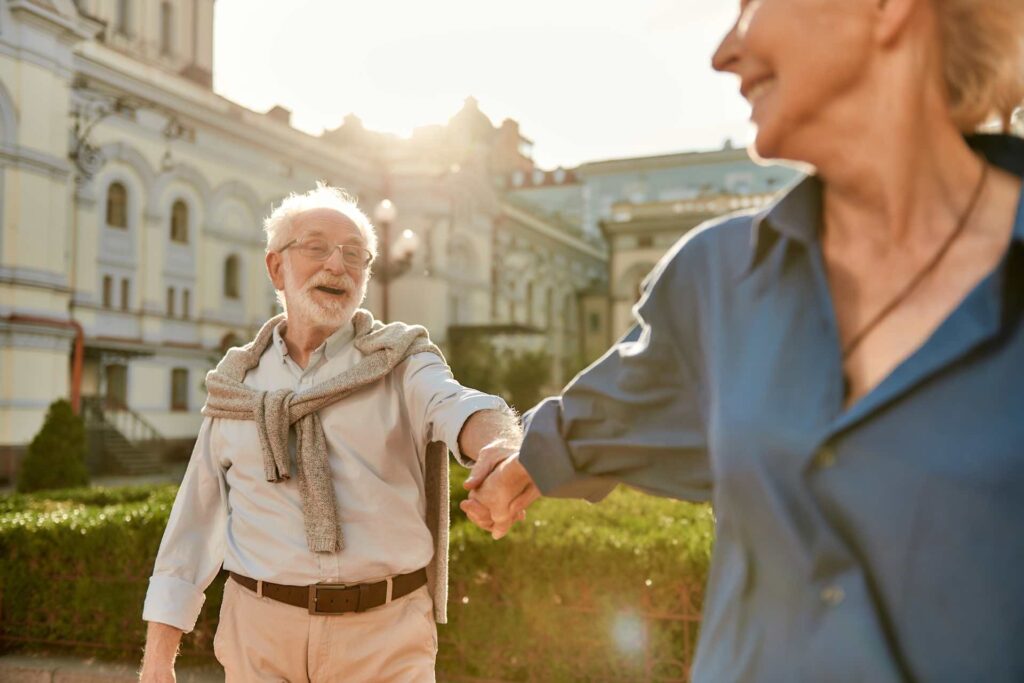Life Health Span positive image. Elderly couple dressed in casual clothes enjoying a walk in the sun among building set in a french arcade. The couple are happy and smiling. Chiropractic treatment to maintain a healthy life.