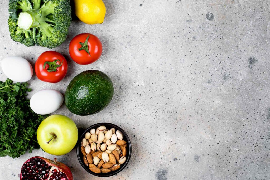 A grey cement table woith fresh fruit and vegetable place a top the table. The fruit has avocado, tomatoes, apples and lemons. The vegetable is a broccoli and egg. Nutrition