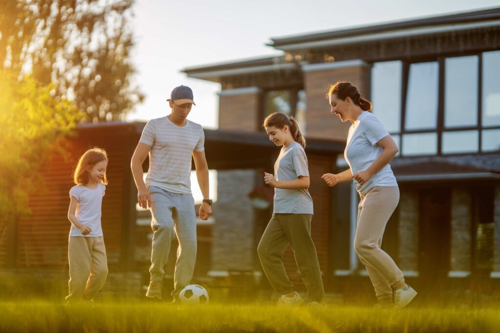 A young family playing soccer outside of there house. The family has a mother, father and two younger kids, both girls. They dressed for autumn type weather with long pants and T shirts