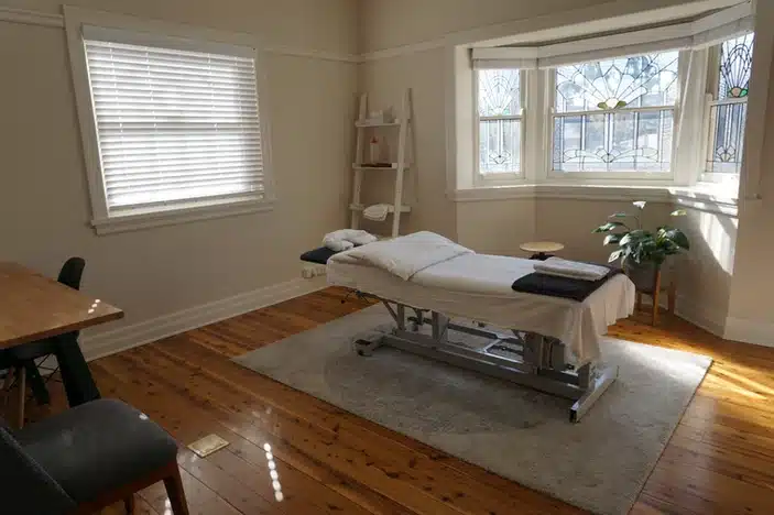 Picture of chiropractic treatment room. A chiropractic table is in the middle of the room on a blue rug. There is a desk and office chair in one corner and a chair for patients to sit on located in another. There is a bay window and the wall colour is cream. A white sink is located in a corner and a plant is located under the window