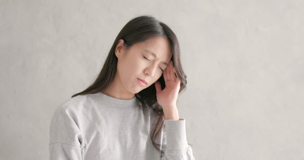 Young Asian female sitting holding the left side of her head with one hand. Representing a Headache. She is wearing a light grey jumper. She has neck pain and a headache.