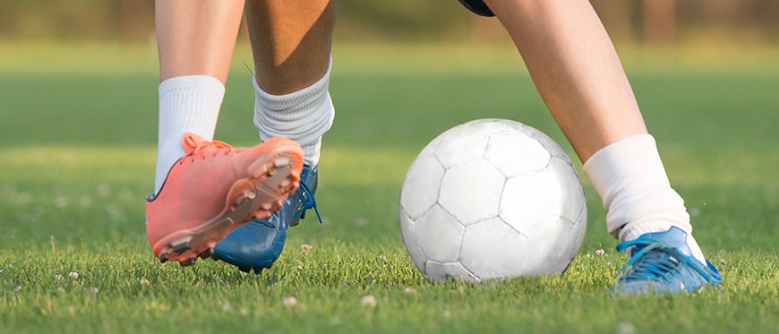 Photo of two young people's lower legs and feet while playing soccer outdoors. both trying to kick a ball. one is wearing orange shoes one is wearing blue shoes.