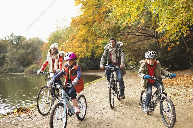 photo of young family riding bikes together outdoors next to a lake or river. there is a young mother and father riding behind 2 young children one boy and one girl, they are all wearing multiple warm layers of clothing. the clothes are green blue and orange. the trees have orange leaves looks like autumn.