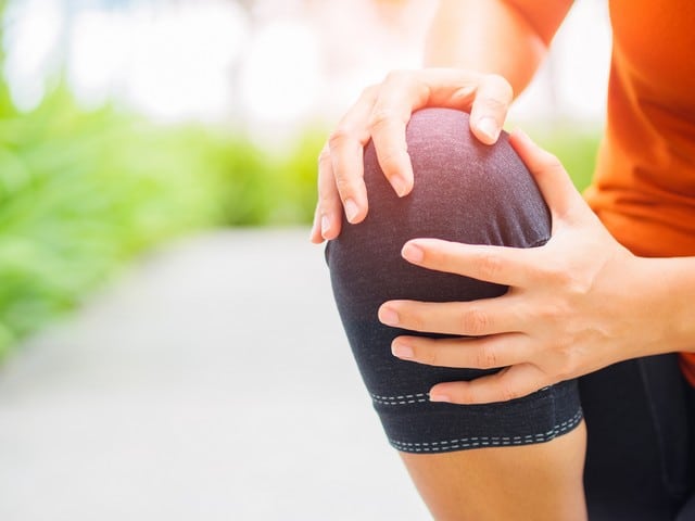 image of a young woman's leg bent with her holding her knee. red highlight over her knee showing she is experiencing pain in her knee. she is wearing grey tights and orange shirt