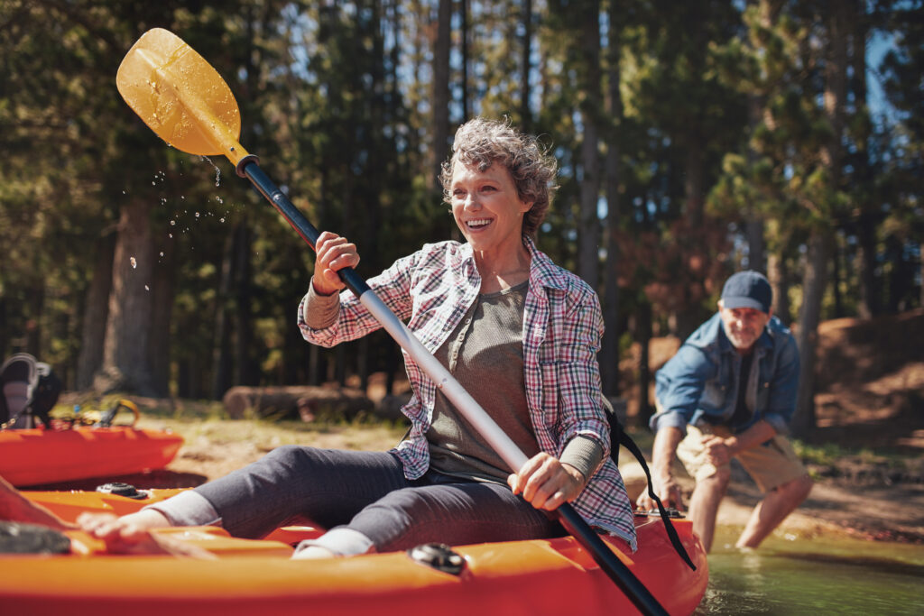 Portrait of happy senior paddling kayak in the lake with man supporting from behind. Mature couple enjoying a day at the lake. The kayak is orange and there are lots of trees in the background