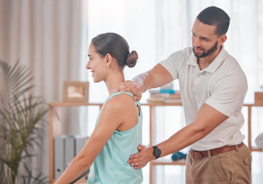 A male osteopathy clinician stands behind a female patient whose wearing a light blue top. The man is palpating the middle of the back.