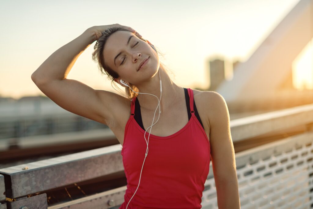 A young lady wearing a red top who is out exercising. She is wearing headphones. She is stretching her head to the right. She is exercising at sunrise.