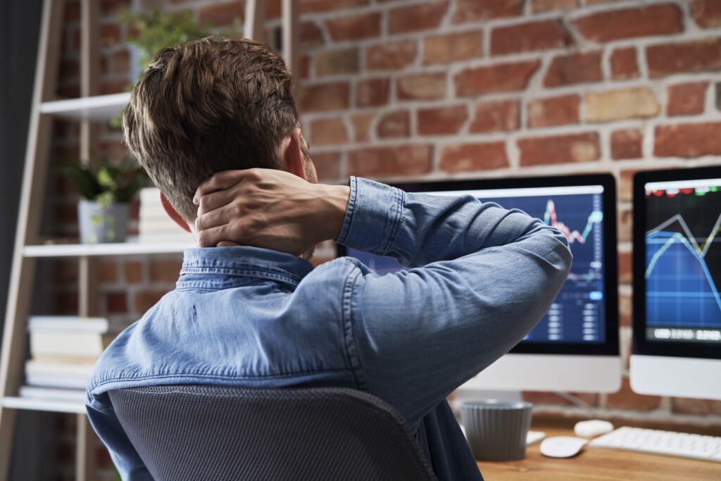 A young man sits at a desk holding his neck in pain. He has compute screens in front of him. He is wearing a blue shirt. neck pain and headaches are represented by hand position.