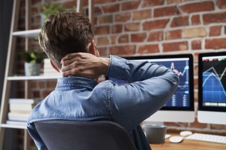 A young man sits at a desk holding his neck in pain. He has compute screens in front of him. He is wearing a blue shirt. neck pain and headaches are represented by hand position. A common neck compliant is shown in this image as many people work at a computer device and develop neck pain.