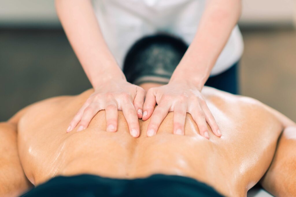 A young male is lying face down, receiving a massage. The clinician is using oil. The benefit of massage is to reduce muscle tension.