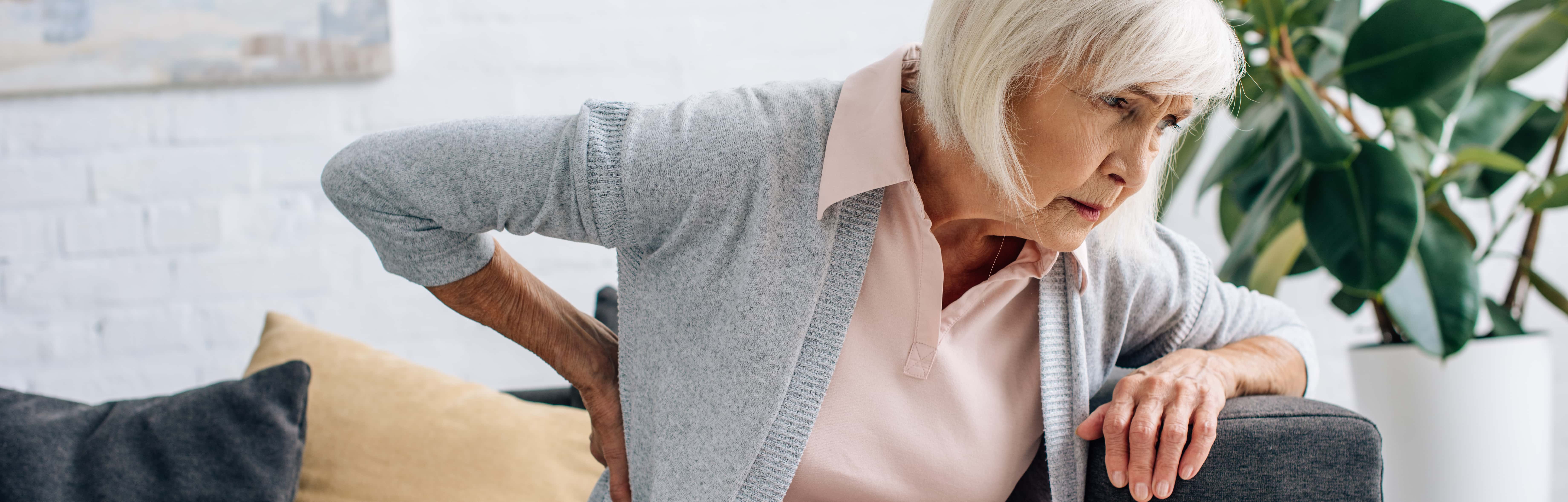 An elderly lady with pain from arthritis. She is wearing a grey sweater. Arthritis pain decreases movement. She looks stiff and in pain. osteoarthritis lumbar pain