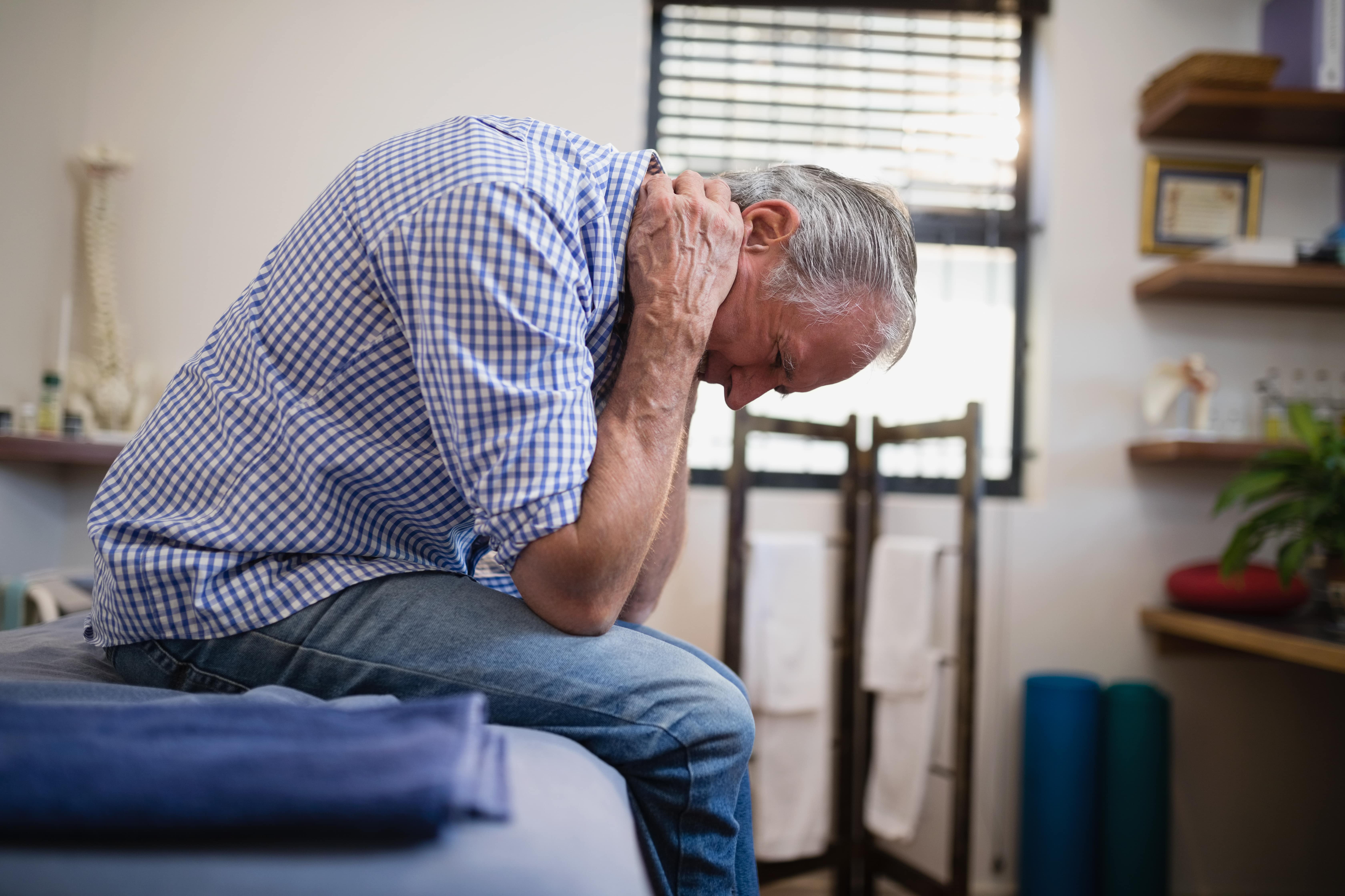 Man sitting on treatment table clutching neck in pain. He is wearing a checked blue shirt. Holding his cervical spine.