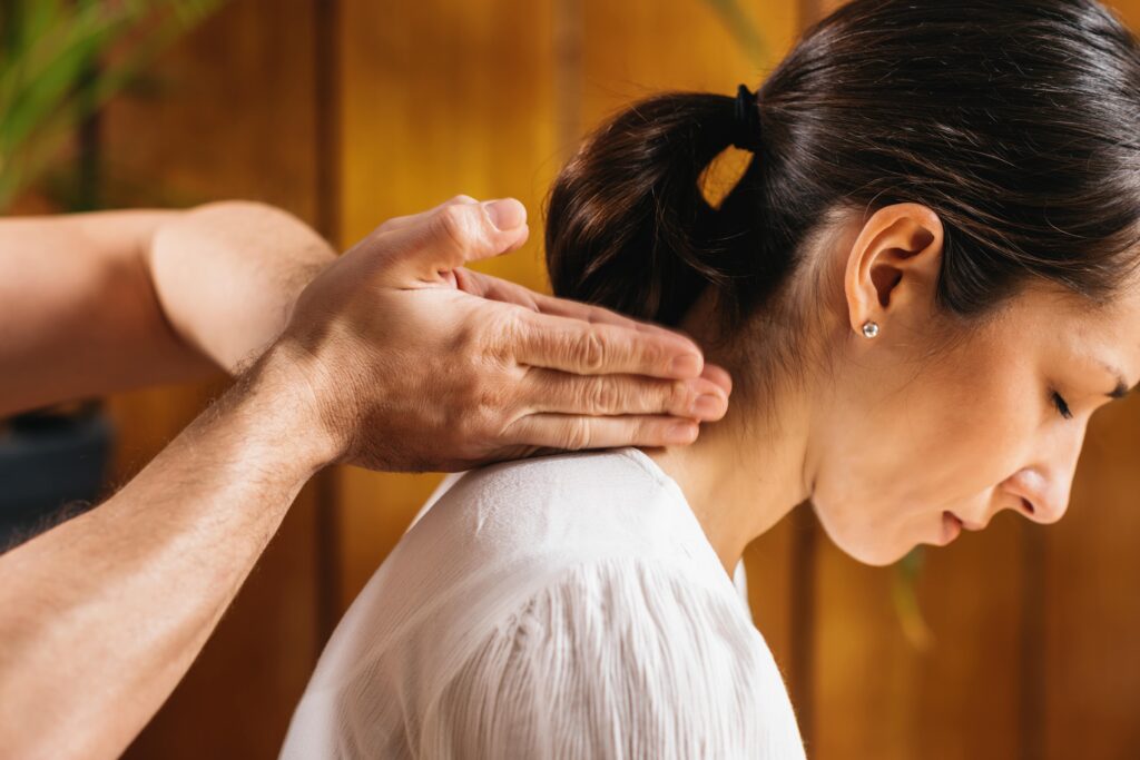 Thai massage Chiswick. Lady is sitting having neck massage for neck pain. You see doctors' hands-on the patients' lower neck. The patient is wearing a white shirt. Massage neck pain.
