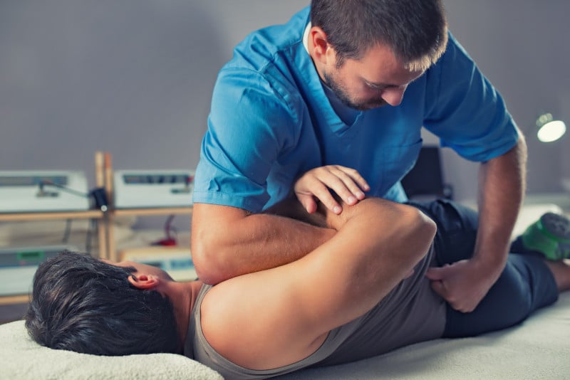 chiropractor treating lower back for back pain. Osteoarthritis and lumbar pain. Patient is lying on the side and chiropractor is rolling him to the left side. Pateint is wearing a grey singlet. Chiroractor is wearing a blue button up shirt.