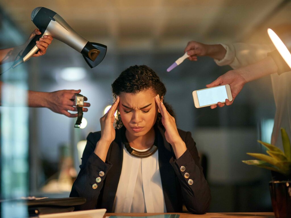 A young lady wearing a black suit jacket with a look of stress on her face as she has phones, hairdryer, watch coming at her from the sides. The picture shows a lady suffering from stress.