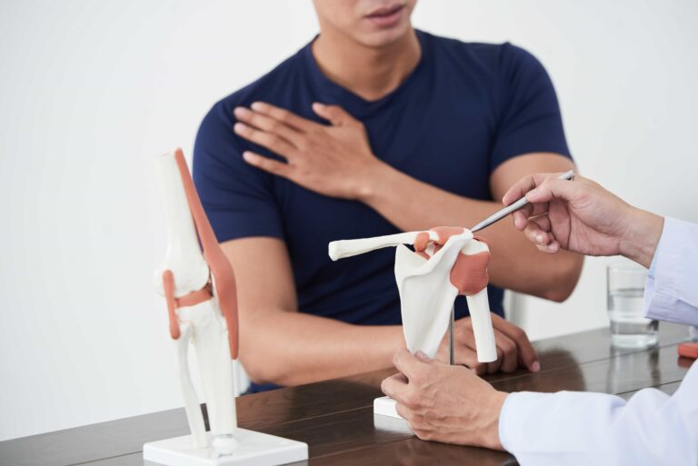 Young man wearing dark blue shirt sitting in a clinic. Doctor is holding model of shoulder anatomy. Rotator cuff muscles are on display. Patient is holding left shoulder showing shoulder pain from rotator cuff injury.