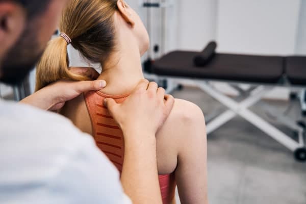 A lady sits on a table in a brown singlet top. Doctor stands behind her performing physical examination of the neck. Chiropractor physical examination.