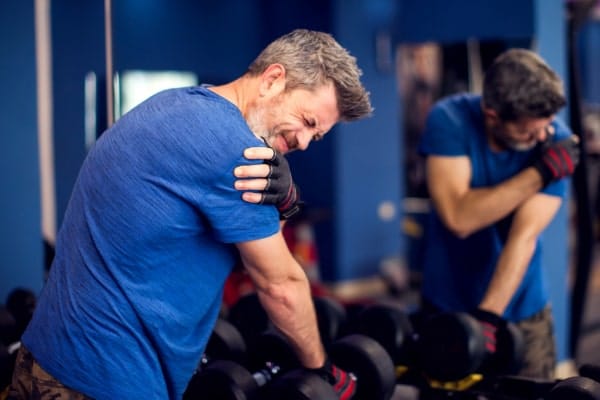 a man wearing a blue T shirt is at the gym, holding a dumbbell. He is holding his right shoulder in pain, showing a rotator cuff injury. He has a painful expression on his face.