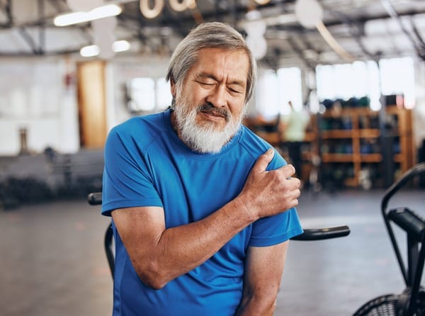 An elderly man is wearing a blue shirt and holding his left shoulder. He has chronic shoulder pain from repetitive use. The background is a wharehouse. Shoulder pain, chronic rotator cuff injury.
