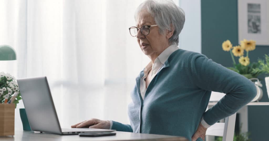 Elderly lady sitting at computer desk, wearinga light blue jumper. Holding her lower back region, showing pain on right side. right side lower back pain