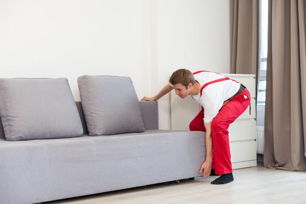 Man wearing red overalls is bending in flexion to lift a lounge. Common cause of lower back pain is his poor posture, with little knee bend.