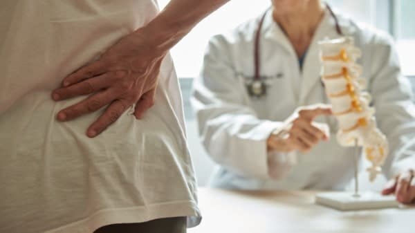 A doctor wearing a white lab coat is explaining nerve compression to a patient from spinal stenosis. Patient is holding lower right side of lumbar back region.