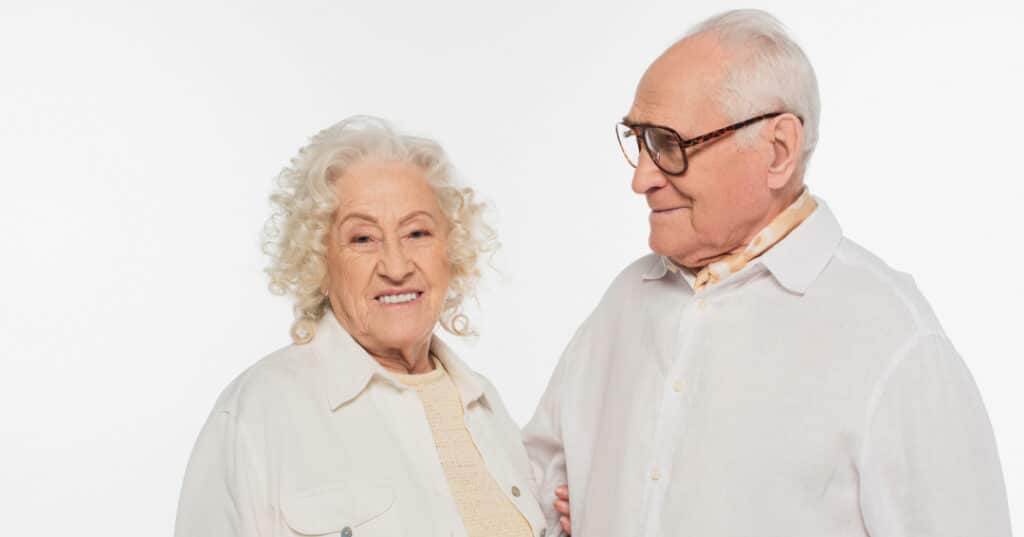 An elderly couple is standing facing each other. The man is wearing glasses.