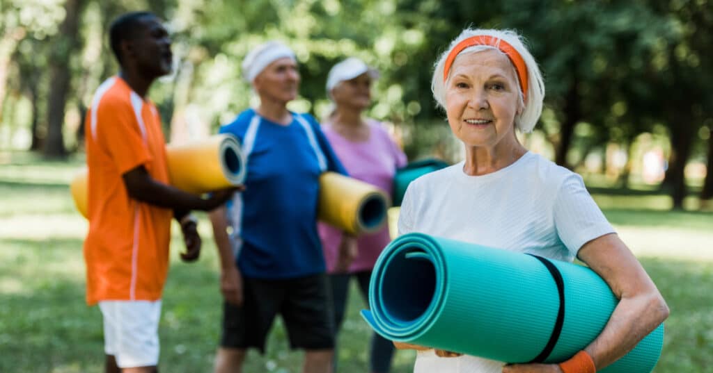 A group of people in a park. One lady is holding a light green yoga mat