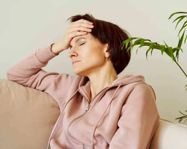 An old lady wearing a pink brown jumper is sitting holding her head, with a headache. She is sitting in a lounge resting against the arm rest. The hand is placed to represent a headache.