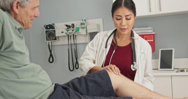 Female surgeon with older male patient in exam room checking range of motion on knee injury.