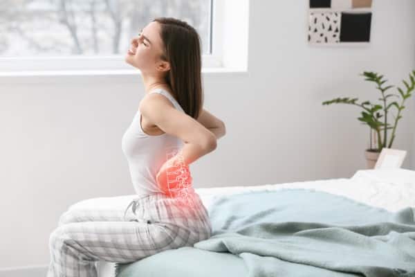 Young woman suffering from back pain caused by facet syndrome. she is sitting on here bed holding her back.