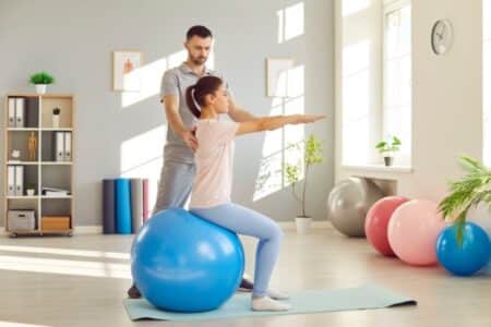 The patient is learning strength and stability movement exercises from a chiropractor and functional movement expert. She is sitting on a blue Swiss ball and wearing a white shirt. The doctor is standing behind her. Retraining your nervous system and proprioception.