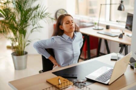 Young businesswoman having backache from sitting at her desk. Her hands are on her back where muscle stiffness and achy pain will occur.