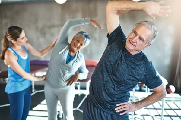 Functional movement of spine and hip for hip spine syndrome. Two elderly patients are standing wearing active wear in a rehab setting. They are reaching up and creating traction in the lumbar spine.