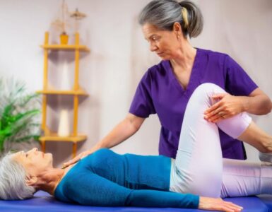 elderly lady lying face up on a treatment table with doctor gently stretching her hip joint. Lady is wearing white pants and blue top.