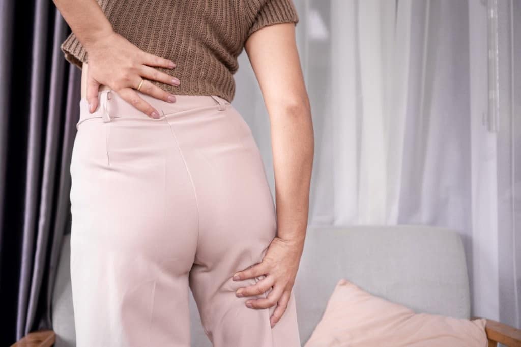 Sciatica related pain is shown by a young lady holding her posterior right leg in pain. She is wearing business attire, with a creamy, brown pant. She is also holding her lower back where pain would be present with sciatica.