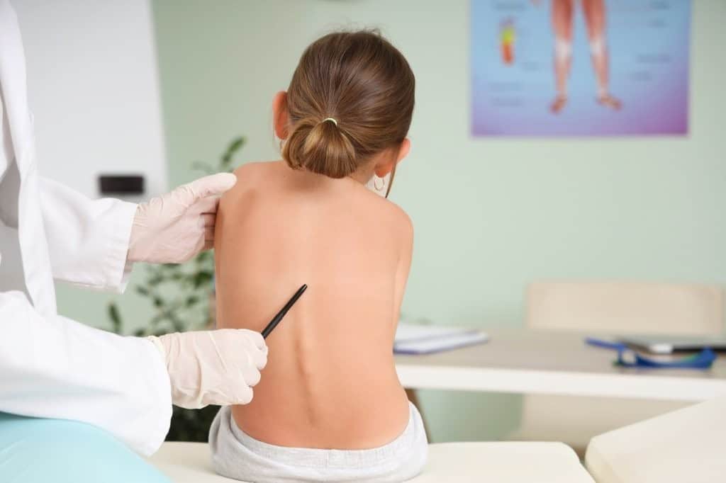 A child is sitting on a treatment table, facing away from the camera. The child has a visible scoliosis to the left side of her lower back.