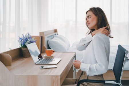 Lady sitting at a desk holding her neck winching in pain. Bad posture leads to neck pain. This picture shows this.