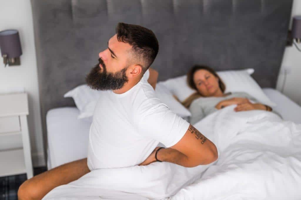 A man sitting on the edge of his bed holding lower back. Person has a strained look of pain on his face. He is wearing a white t-shirt.