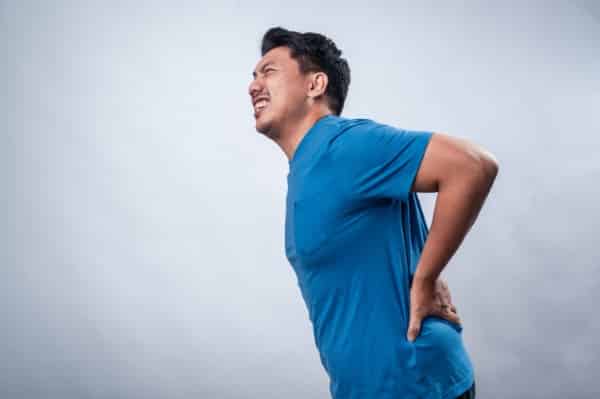 An Asian man wearing a blue t-shirt against a white isolated background is holding his lower back with a pained expression, indicating back pain or discomfort. His bent posture suggest strain